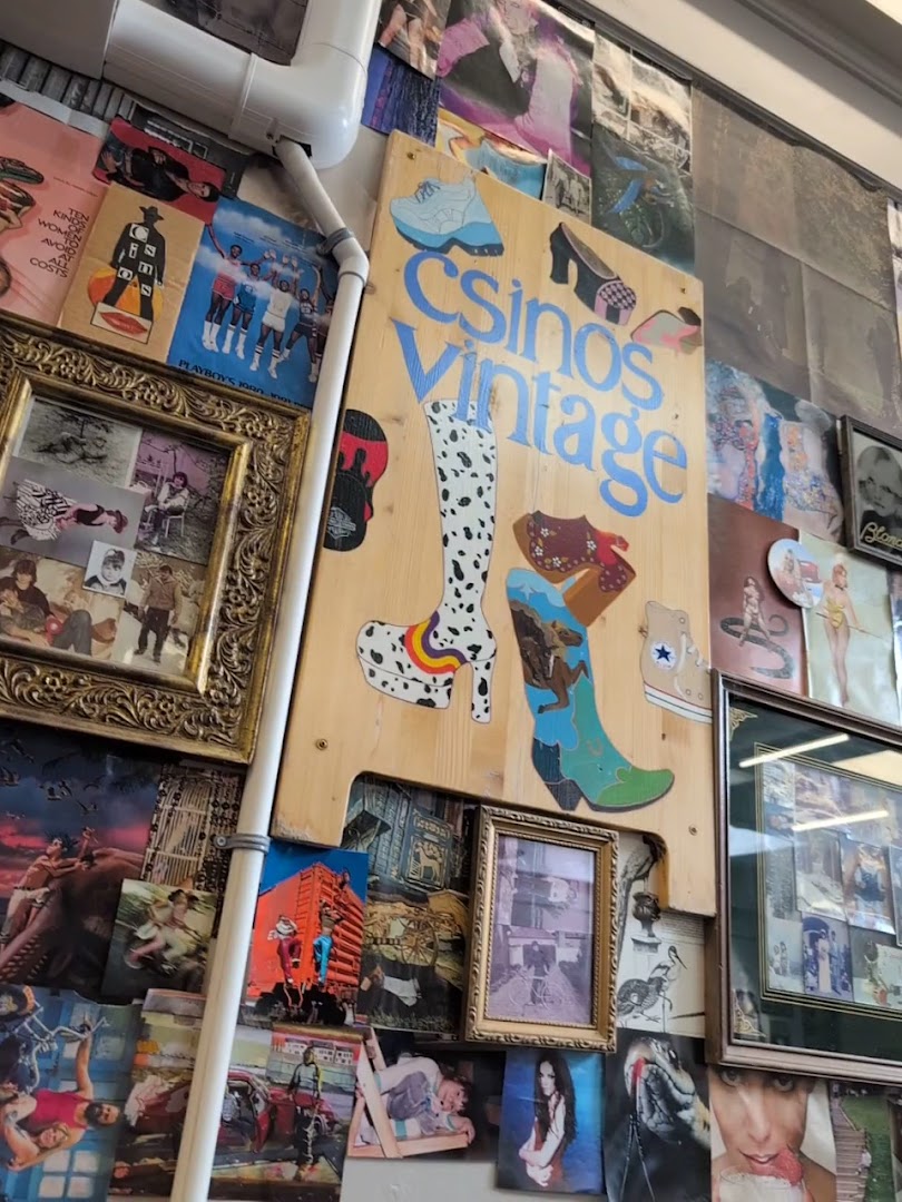A hand-painted 'Csinos Vintage' sign featuring illustrated boots and heels, mounted on a wall dense with posters, framed photos, and vintage ephemera.