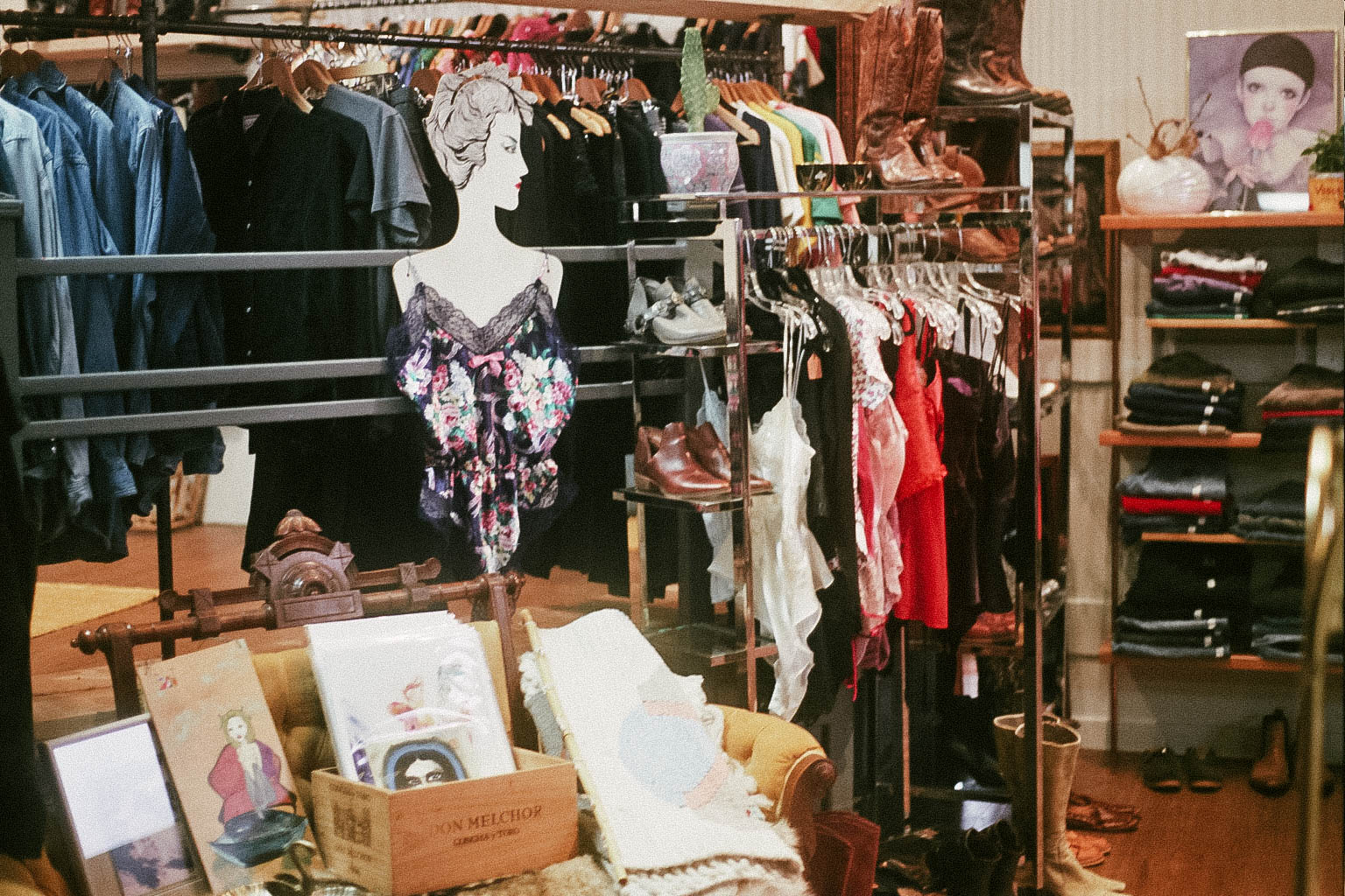 A rack of leather jackets and dark clothing beside a mannequin torso wearing a floral lace slip dress, with a glass shelf of shoes and handbags and a wine crate of framed art below.