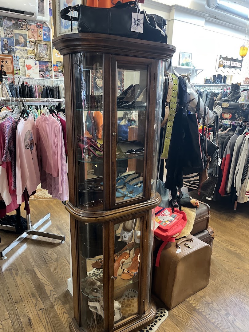 A wooden curio cabinet displaying shoes and accessories beside a 'Star Vintage' sign, with belts, bags, and vintage suitcases nearby.