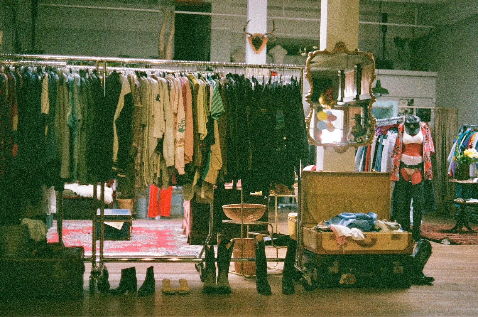 A dense clothing section reflected in an ornate gold-framed mirror, with a red patterned rug and a mannequin dressed in denim visible in the background.