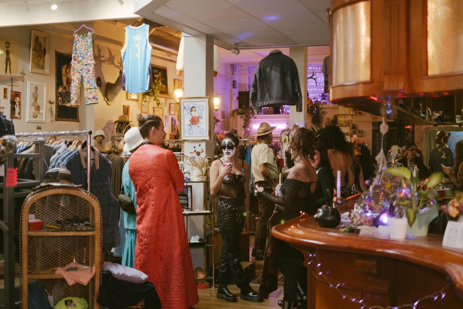 A lively night inside the shop under purple lighting, with shoppers browsing racks near the bar counter and a framed Csinos Vintage print on the pillar.